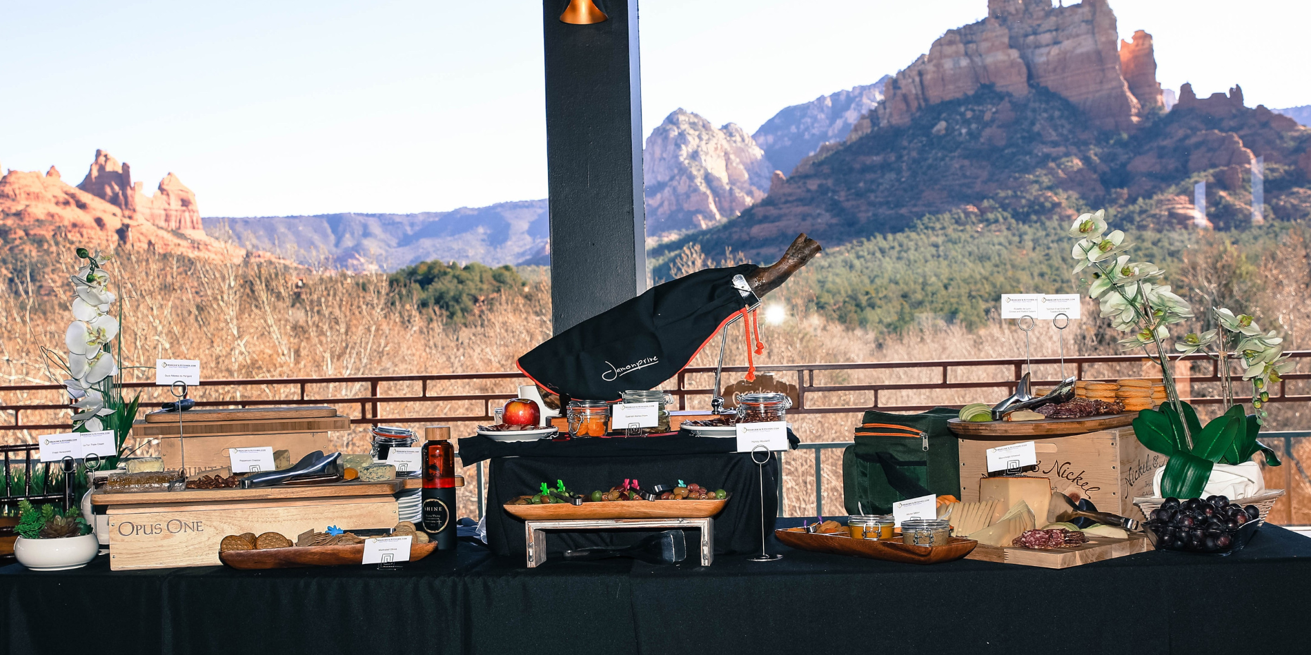 This image showcases a gourmet catering spread on a buffet table at an indoor event at the view at creekside with the majestic red rocks of Sedona in the background. The table is adorned with a variety of cheeses, charcuterie, and fruits, elegantly presented in wooden crates and on rustic boards. A notable feature is a large bottle of Opus One wine in a bespoke cradle with a black cloth, indicating a high-end dining experience. The setup is complemented by green floral arrangements and placards describing the wine selections, all under the open sky, inviting guests to enjoy a luxurious culinary experience in a stunning natural setting.